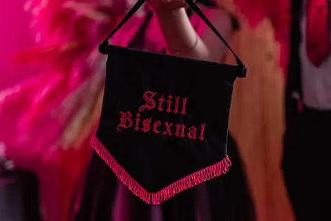 A black banner with pink fringing along the bottom with 'still bisexual' written in pink in the centre