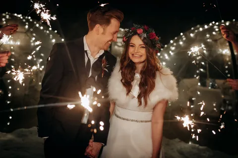 Bride and groom holding hands, with big smiles on their faces during their sparklers exit. Bride is wearing a flower headband of green and red, a white fur shawl and a white wedding dress, and the groom is wearing a dark suit with an undone bowtie