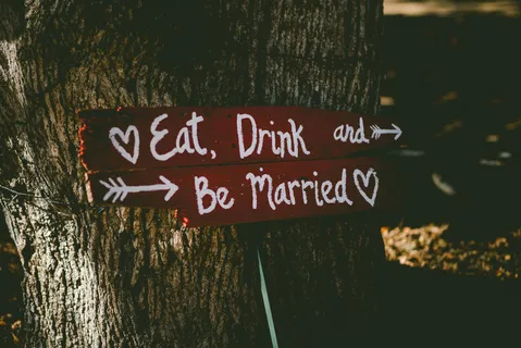 A handwritten sign saying 'eat, drink & be married' with hearts and arrows, attached to a tree